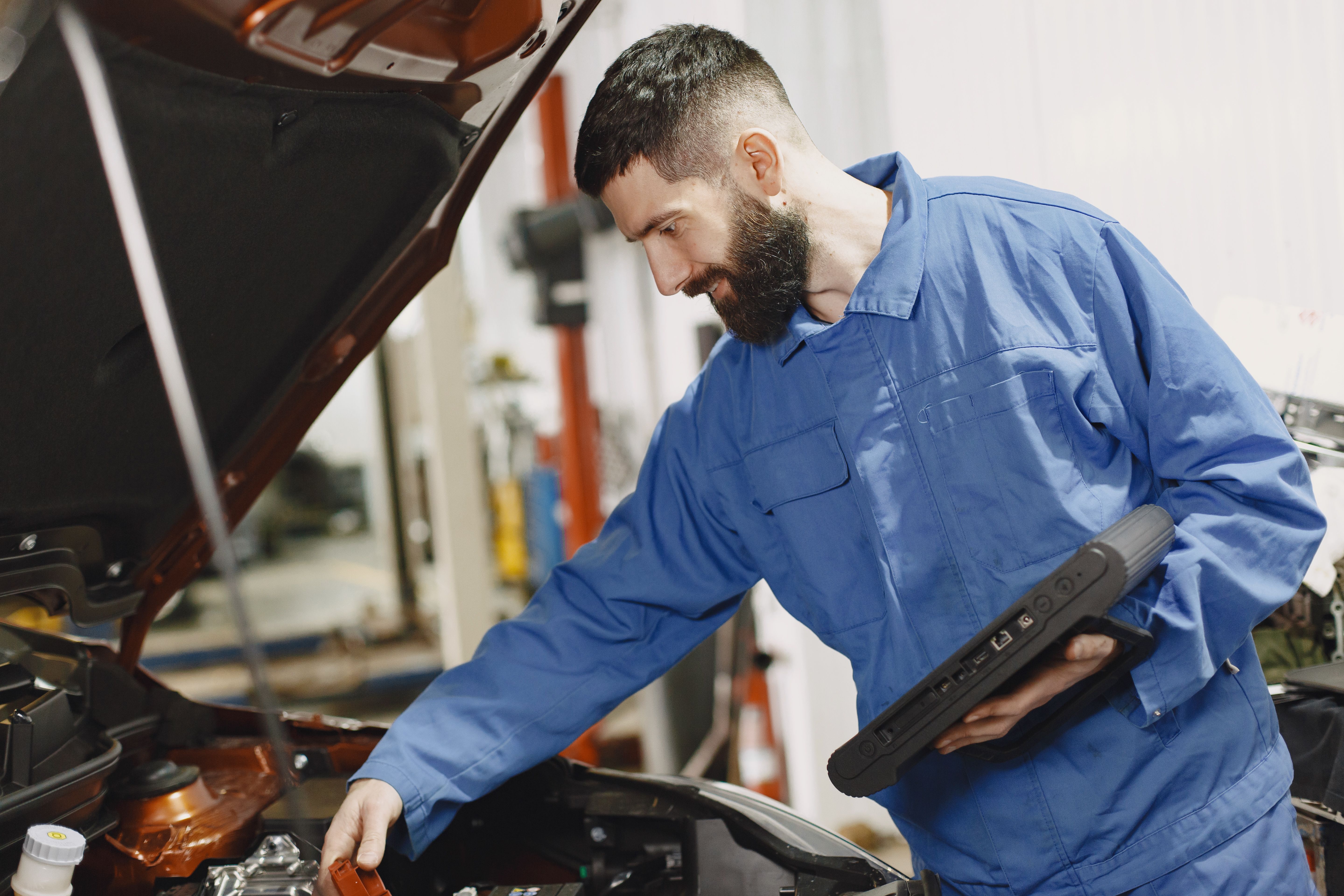 Technician in blue coveralls using a rugged diagnostic tablet while inspecting a vehicle engine bay